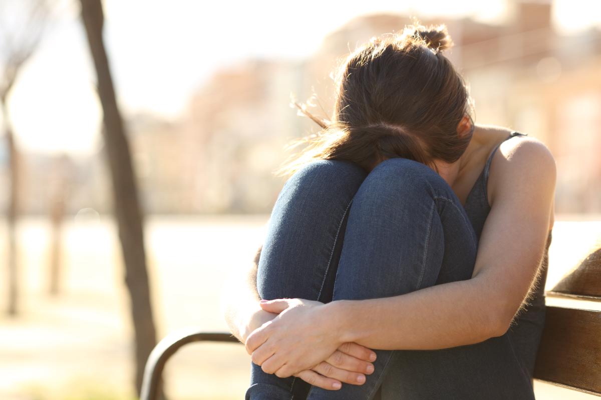 a girl sitting with her knees to her face on a bench thinking about signs of teen self-harm