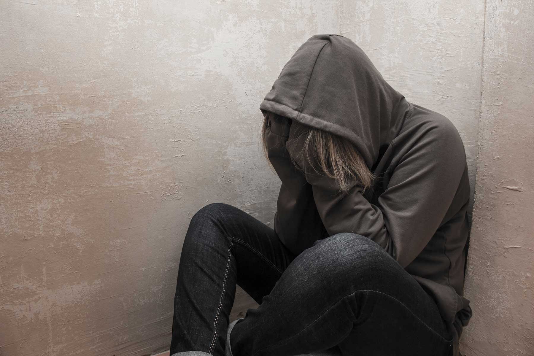 Person sitting in a corner wearing a hoodie and covering their face with their hands while experiencing symptoms of drug withdrawal